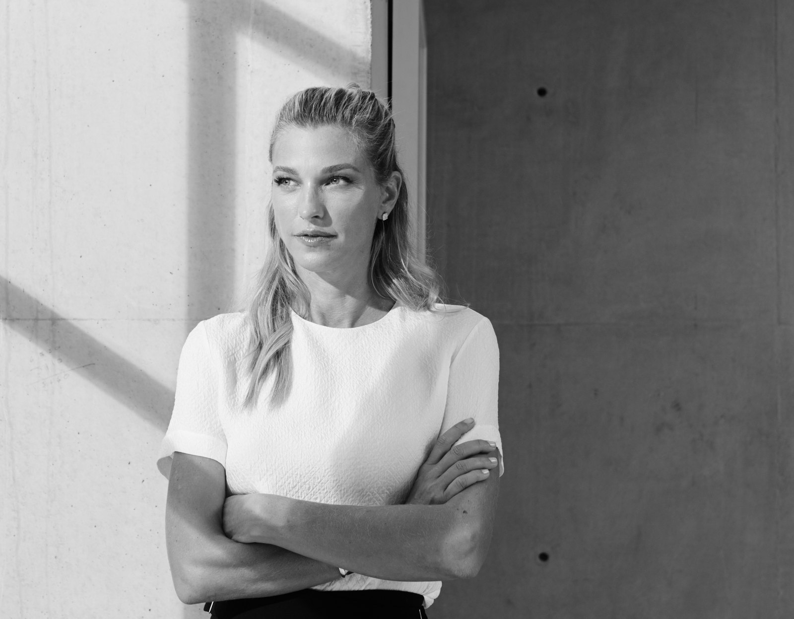 Nicole Junkermann in un ritratto editoriale in bianco e nero, con le braccia conserte accanto a una parete di cemento illuminata dal sole